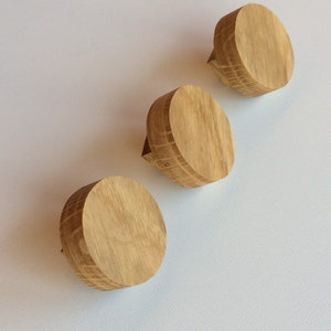 Puede incluir: Tres pomos de madera para armarios con acabado de madera natural dispuestos en diagonal sobre un fondo blanco. Cada pomo es circular con una peque&ntilde;a proyecci&oacute;n rectangular en un lado.