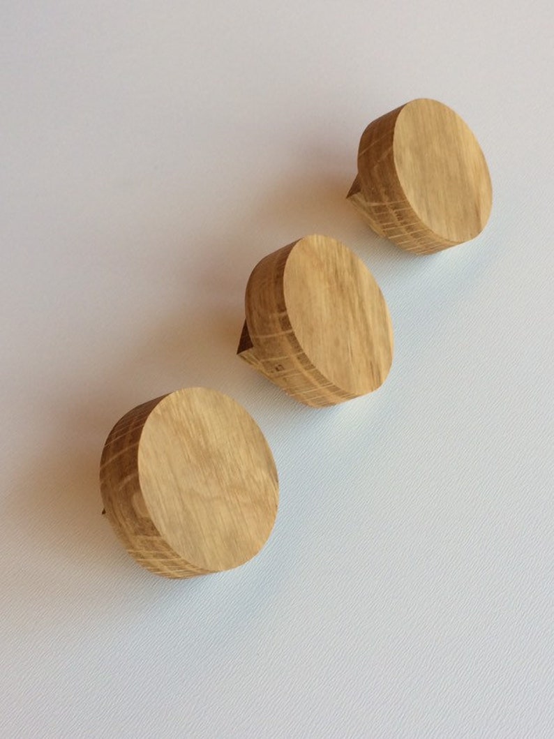 Puede incluir: Tres pomos de madera para armarios con acabado de madera natural dispuestos en diagonal sobre un fondo blanco. Cada pomo es circular con una peque&ntilde;a proyecci&oacute;n rectangular en un lado.