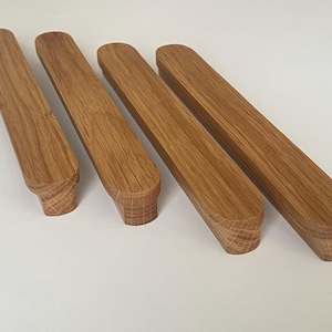 Op de afbeelding: Vier houten kastgrepen met een natuurlijke eiken afwerking. De rechthoekige grepen hebben afgeronde uiteinden en een decoratieve basis. De houtnerf is zichtbaar. Ze zijn gerangschikt op een witte ondergrond.