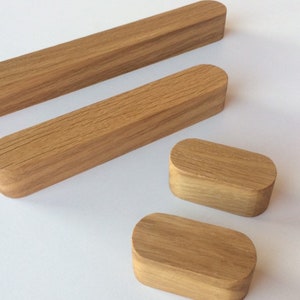 May include: Light brown wooden cabinet pulls and knobs. The pulls are long and rectangular with rounded ends, while the knobs are small, rectangular blocks. The wood grain is visible on the surface of the pulls and knobs.