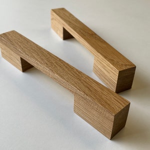 Può includere: Due maniglie per mobili in legno con finitura in rovere naturale. Ogni maniglia ha una barra rettangolare con un blocco quadrato ad ogni estremità e un ritaglio rettangolare al centro. Le maniglie sono su uno sfondo bianco.