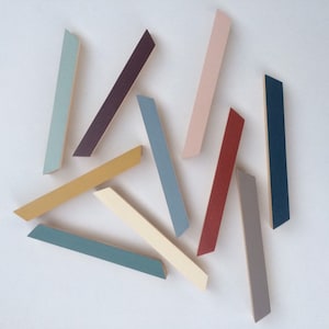 May include: An overhead shot of ten colorful, angled wooden pieces. The pieces are painted in various colors, including blue, yellow, red, and gray. The pieces are arranged on a white surface.