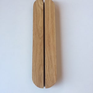 Puede incluir: Dos tiradores de puerta verticales de madera clara. Cada tirador tiene una parte superior redondeada y una ranura vertical en el centro. Los tiradores est&aacute;n montados en una pared blanca.