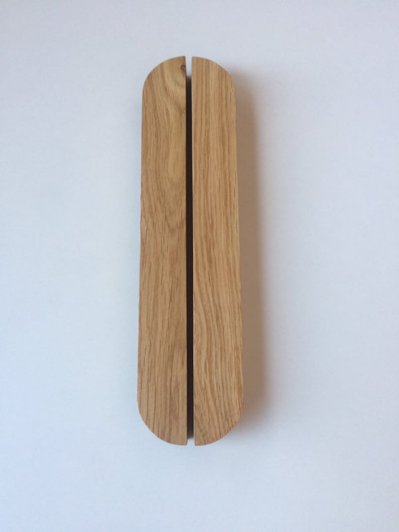 Puede incluir: Dos tiradores de puerta verticales de madera clara. Cada tirador tiene una parte superior redondeada y una ranura vertical en el centro. Los tiradores est&aacute;n montados en una pared blanca.