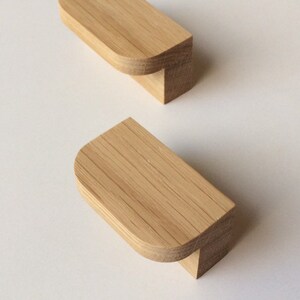 Può includere: Due pomelli per mobili in legno con finitura naturale. Ogni pomello ha una parte superiore rettangolare con bordi arrotondati e una piccola base quadrata. La venatura del legno è visibile, su sfondo bianco.