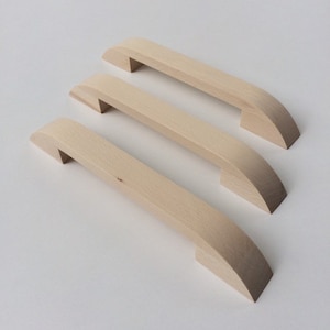 Può includere: Tre maniglie per mobili in legno non rifinite. Ogni maniglia ha un corpo rettangolare con estremità curve e una piccola sporgenza rettangolare su ciascuna estremità. Il legno è di colore chiaro naturale.