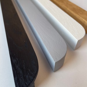 Può includere: Primo piano di quattro binari di cornice in legno di vari colori. I colori includono nero, grigio, bianco e legno naturale. I binari hanno un bordo arrotondato e sono disposti fianco a fianco, mostrando le diverse finiture disponibili per l'incorniciatura di opere d'arte.