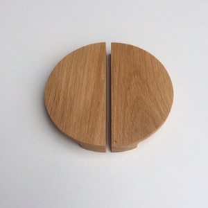 May include: Two matching wooden cabinet pulls, each a half-circle shape, are displayed against a white background. The pulls are made of light-colored wood, possibly oak, and have a natural grain pattern. They are designed to be mounted on a cabinet door.