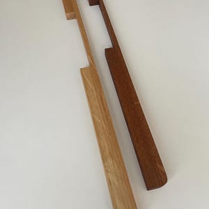 Könnte beinhalten: Zwei Holzschrankgriffe. Einer ist hell eichenfarben, der andere dunkelbraun. Beide haben einen rechteckigen Griff mit einem Ausschnitt und einen langen, schmalen Körper. Die Griffe sind vor einem weißen Hintergrund platziert.