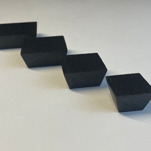 Könnte beinhalten: Vier schwarze, geometrische Holzblöcke diagonal angeordnet. Jeder Block hat eine quadratische Oberseite und eine trapezförmige Form mit sichtbarer Holzmaserung. Die Blöcke sind gleichmäßig vor einem weißen Hintergrund angeordnet und erzeugen Tiefe und Ordnung.
