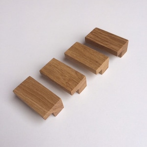 Può includere: Quattro piccoli blocchi di legno rettangolari con finitura a venatura naturale. Ogni blocco ha un taglio angolato unico su un'estremità e un'intaccatura sull'altra, suggerendo un possibile design ad incastro. I blocchi sono disposti diagonalmente su una superficie bianca.