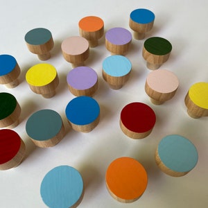 Peut inclure: Ensemble de boutons de meubles en bois avec une base en bois naturel et des dessus circulaires peints de différentes couleurs, dont le bleu, l'orange, le vert et le rose. Les boutons sont disposés sur une surface blanche, mettant en valeur leurs teintes et leur design.