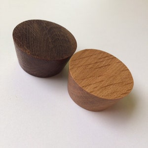 Può includere: Due tappi per bottiglie in legno. Uno è marrone scuro, l'altro è marrone chiaro. Entrambi hanno una forma cilindrica con la parte superiore inclinata. La venatura del legno è visibile su entrambi i tappi. Su sfondo bianco.