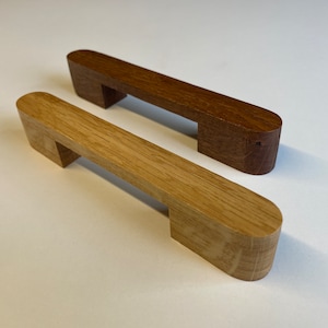 Può includere: Due maniglie per mobili in legno, una in rovere chiaro e una in noce scuro, sono esposte su una superficie bianca. Ogni maniglia ha una barra rettangolare con estremità arrotondate e due piedi quadrati per il montaggio.