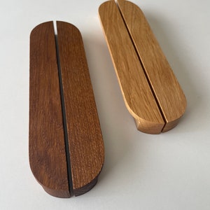 May include: Two wooden phone stands. One is a dark brown colour, and the other is a light brown colour. Both stands have a curved top and a slot down the middle to hold a phone. The stands are made of wood.