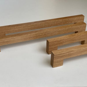 Può includere: Una collezione di quattro maniglie per mobili in legno di varie dimensioni. Le maniglie sono rettangolari con un'area di presa incassata e sono realizzate in legno marrone chiaro. La venatura del legno è visibile. Le maniglie sono disposte su una superficie bianca.