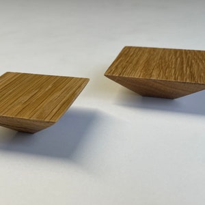 Può includere: Due oggetti in legno a forma di diamante, di colore marrone chiaro e con venature visibili. Gli oggetti hanno un bordo smussato, creando un design geometrico. Sembrano piccoli oggetti decorativi o componenti.