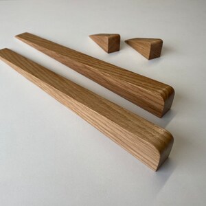 Puede incluir: Dos barras largas de madera marrón claro con bordes angulados y extremos redondeados, junto con dos pequeñas piezas triangulares de madera. La veta de la madera es visible. Artículos para decoración o mobiliario.