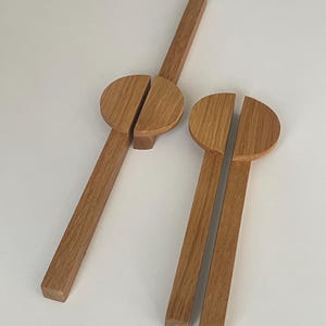 Könnte beinhalten: Zwei Salatbestecke aus Holz. Jedes Besteck hat einen langen, rechteckigen Griff und einen geteilten, halbkreisförmigen Kopf. Das Holz hat eine natürliche, hellbraune Farbe. Das Besteck ist auf einer weißen Oberfläche angeordnet.