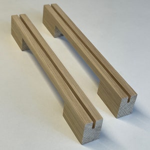 Könnte beinhalten: Zwei hellfarbige Holzschrankgriffe. Jeder Griff hat einen rechteckigen Sockel mit einem vertikalen Schlitz und einen langen, rechteckigen Griff mit einer Nut. Die Holzmaserung ist sichtbar. Diese Griffe sind für Schränke oder Schubladen konzipiert.