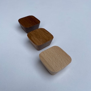Peut inclure: Trois boutons de meuble en bois de différentes nuances de marron. Les boutons sont carrés avec des coins arrondis et une base effilée. Le grain du bois est visible, mettant en valeur la texture naturelle. Chaque bouton mesure environ 3 cm de large.