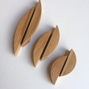 Puede incluir: Tres tiradores de armario de madera con forma de hoja. Cada tirador está hecho de madera de color claro y presenta un diseño dividido. Los tiradores están dispuestos sobre una superficie blanca, mostrando su veta natural y forma única.
