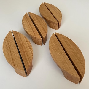 Puede incluir: Cuatro soportes de madera con forma de hoja, con una ranura vertical en el centro. Los soportes son de madera marrón claro y están dispuestos sobre una superficie blanca. Probablemente para exhibir fotos o tarjetas.