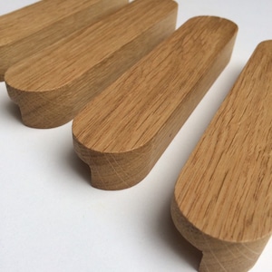 Puede incluir: Cuatro tiradores de armario de madera de roble con acabado natural. Cada tirador es rectangular con bordes redondeados y una hendidura curva en un extremo. Están dispuestos en diagonal sobre un fondo blanco.
