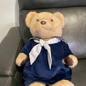 May include: A plush teddy bear wearing a blue velvet dress with a white sailor collar and bow. The bear has brown eyes and a brown nose.