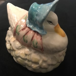 May include: A white ceramic figurine of a duck wearing a blue bonnet and a pink dress with green floral accents. The duck is sitting on a nest of straw.