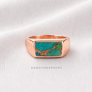 May include: A rose gold ring featuring a rectangular turquoise stone inlay. The turquoise stone displays a mix of blue and green shades with brown matrix patterns. The ring is set against a white fabric background. The text "GEMSTONEPOINT" is visible.