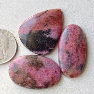 May include: Three pink and black rhodochrosite cabochons, two oval and one teardrop shaped, with a US quarter for size comparison.