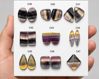 Puede incluir: Una colección de nueve pares de piedras preciosas de fluorita pulida dispuestas en un tablero blanco, etiquetadas de C39 a C47. Cada par presenta patrones de bandas distintos en tonos morado, amarillo y gris, con varias formas geométricas.