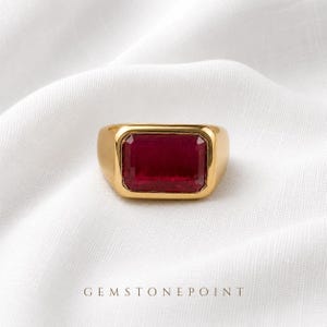 Op de afbeelding: Een goudkleurige ring met een grote, rechthoekige, donkerrode edelsteen gezet in een gladde, gepolijste metalen band. De tekst GEMSTONEPOINT is in kleine, donkere hoofdletters direct onder de ring op de witte stof gedrukt.