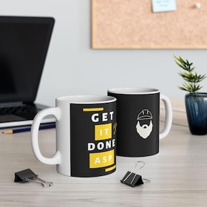 May include: Two white ceramic mugs. One mug has the text "GET IT DONE ASP" in yellow and white on a black background. The other mug has a white illustration of a bearded man wearing a hard hat on a black background.