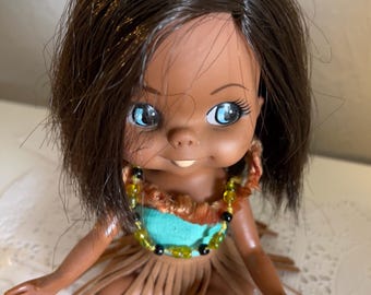 May include: A small doll with brown hair and blue eyes wearing a turquoise top and a brown fringed skirt. The doll is wearing a necklace with green and black beads.
