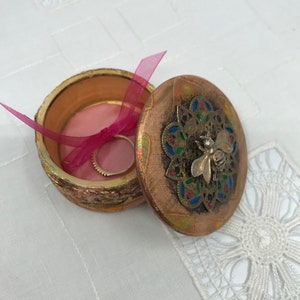 Può includere: Una piccola scatola per anelli rotonda di colore dorato con un interno rosa e un design di ape sul coperchio. Il coperchio è decorato con un motivo a mandala in oro, blu, verde e rosso.