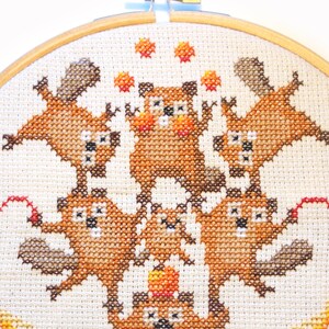 May include: Cross-stitch embroidery hoop featuring a whimsical design of stacked brown squirrels juggling orange balls. The squirrels are arranged in a pyramid shape with a red ball at the bottom. The background is a cream-colored fabric with a decorative orange and yellow design.