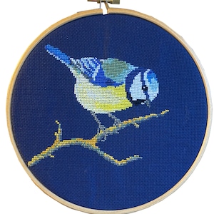 Puede incluir: Bordado de punto de cruz de un herrerillo común posado en una rama, sobre un fondo azul oscuro. El ave presenta detalles azules, amarillos y blancos. El bordado está enmarcado en un aro de madera.