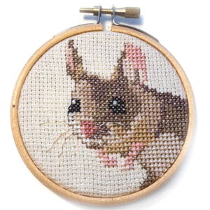 May include: A close-up of a brown and white embroidered mouse in a wooden embroidery hoop. The mouse has pink ears and a pink nose.