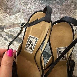 Puede incluir: Un par de sandalias de tac&oacute;n alto a cuadros blancos y negros con correas negras. Las sandalias tienen una plantilla dorada y la etiqueta "GIANNI VERSACE" es visible en la plantilla. Las sandalias est&aacute;n sobre una superficie estampada.