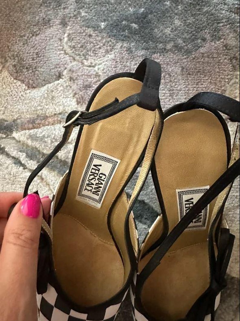 Puede incluir: Un par de sandalias de tac&oacute;n alto a cuadros blancos y negros con correas negras. Las sandalias tienen una plantilla dorada y la etiqueta "GIANNI VERSACE" es visible en la plantilla. Las sandalias est&aacute;n sobre una superficie estampada.