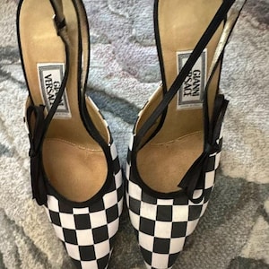 Puede incluir: Un par de zapatos de tac&oacute;n alto a cuadros blancos y negros con punta. Los zapatos tienen correas y lazos negros, y la plantilla presenta una etiqueta que dice "GIANNI VERSACE".