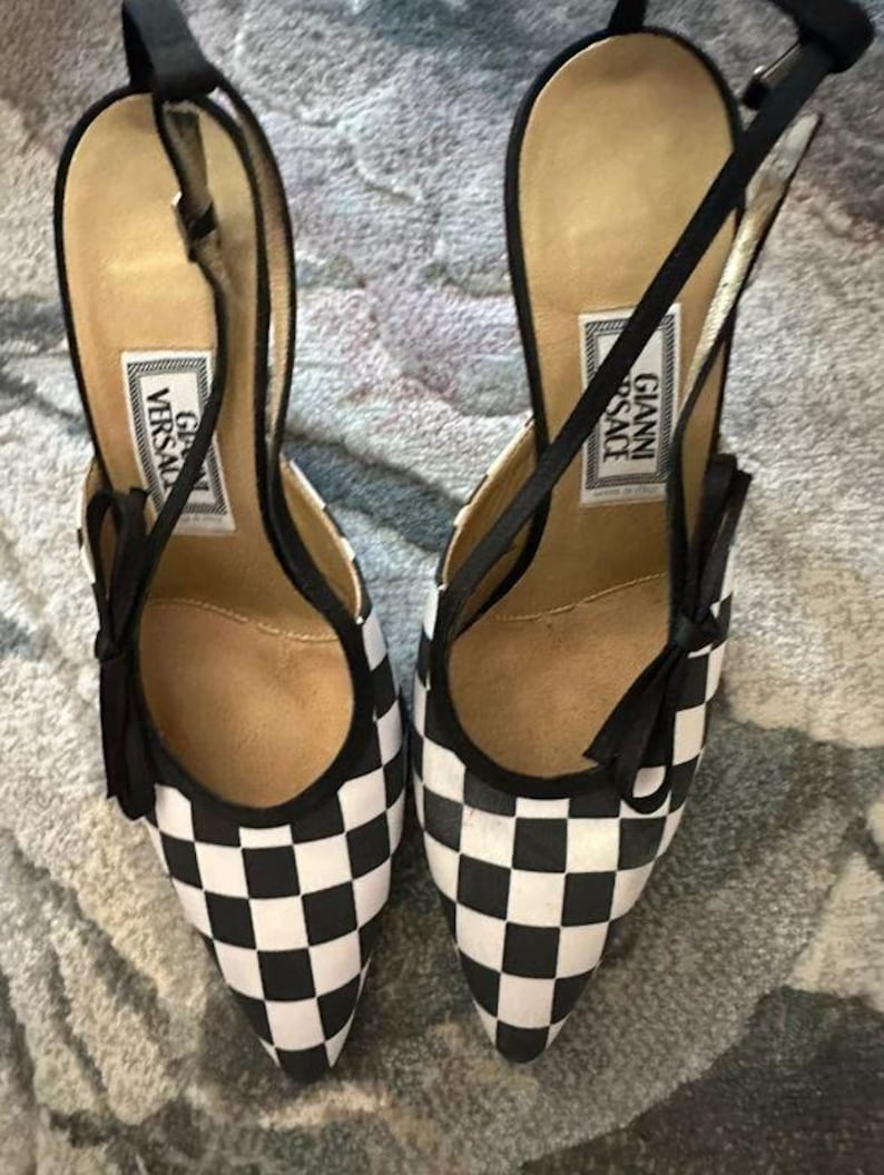 Puede incluir: Un par de zapatos de tac&oacute;n alto a cuadros blancos y negros con punta. Los zapatos tienen correas y lazos negros, y la plantilla presenta una etiqueta que dice "GIANNI VERSACE".