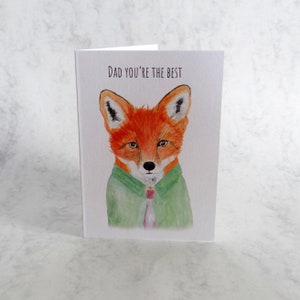 May include: A greeting card with a watercolour illustration of a fox in a green jacket and pink tie. The text "DAD YOU'RE THE BEST" is printed at the top. The fox has orange fur and a white face, with a smart appearance.