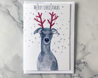 May include: A watercolor Christmas card featuring a greyhound dog wearing red reindeer antlers and a green headband. The words "MERRY CHRISTMAS" are printed at the top of the card. The background has a white base with grey speckles.