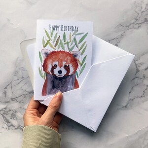 May include: A white greeting card with a watercolor illustration of a red panda, with the words "HAPPY BIRTHDAY" at the top. The card is inside a white envelope.