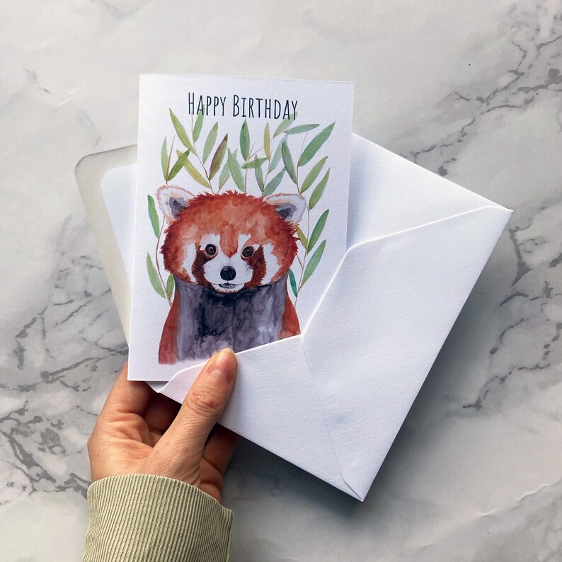 May include: A white greeting card with a watercolor illustration of a red panda, with the words "HAPPY BIRTHDAY" at the top. The card is inside a white envelope.
