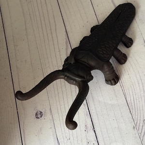 May include: A dark brown cast iron boot pull shaped like an alligator. The boot pull has a detailed textured design and a curved handle for leverage. The alligator's body is the main part of the boot pull, with the handle extending from the front.