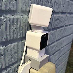 May include: A white security camera system mounted on a blue brick wall. The system includes a camera, a power adapter, and a protective cover. The camera has a black lens and is designed for outdoor use.
